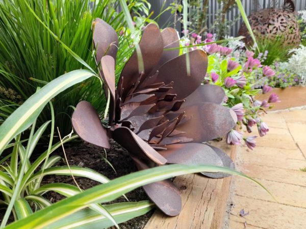 Rusted metal flower sculpture made from Corten steel, featuring a rich orange-red patina, set in a garden. The sculpture measures 36cm in diameter and adds rustic charm to outdoor spaces.